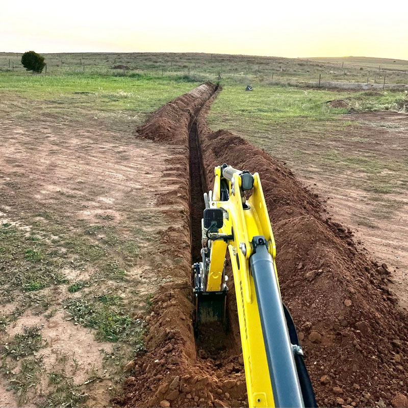 Excavator digging a long, narrow trench across an open paddock