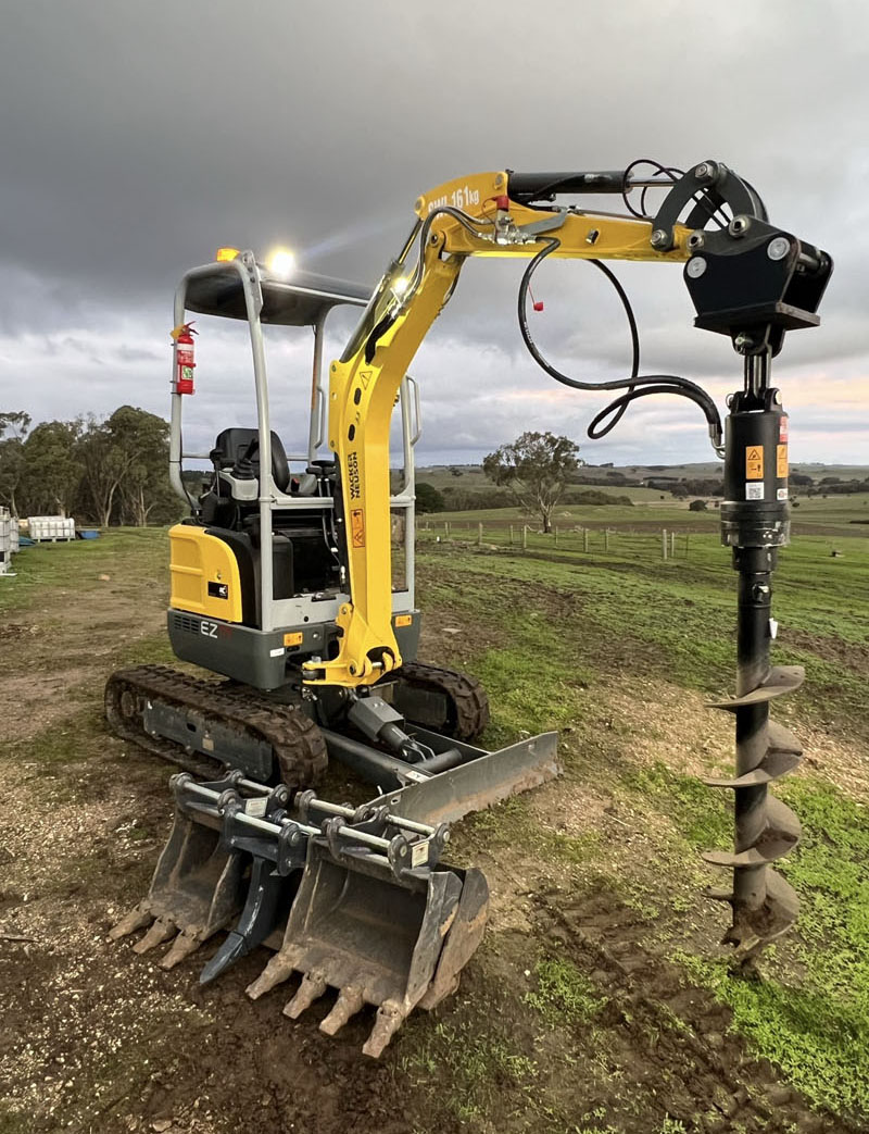 Mini excavator with an auger fitted and spare buckets in front, in a paddock