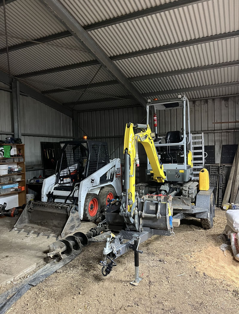Mini excavator on its trailer beside a skid steer loader in a machinery shed