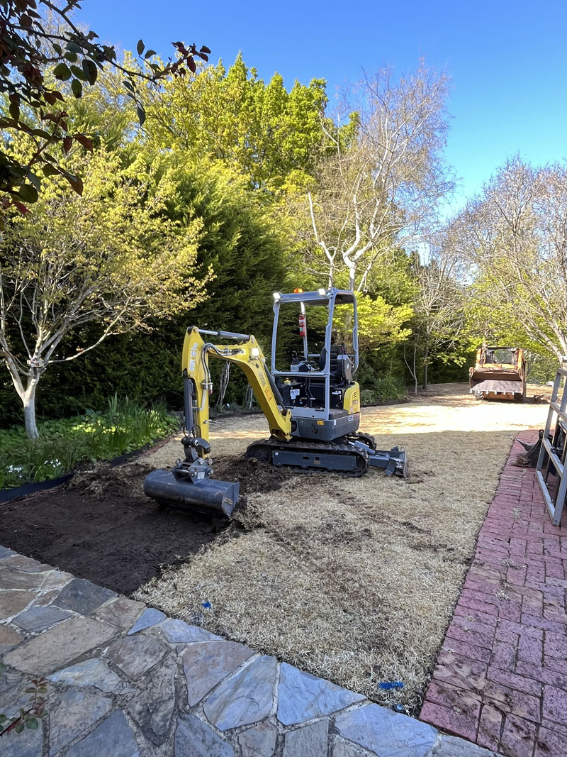 Mini excavator on a levelled sandy base between a brick path and garden beds