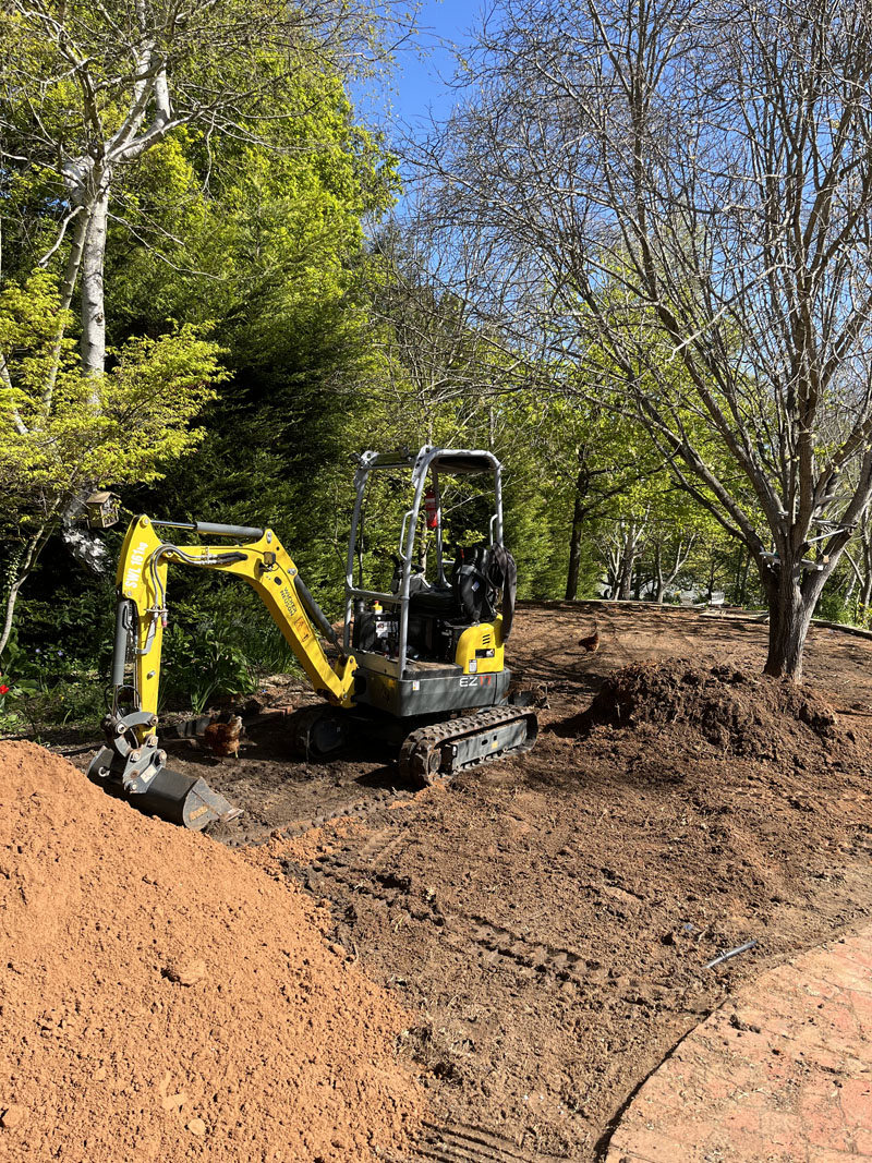 Mini excavator turning over a garden bed under tall trees, with a soil pile in front