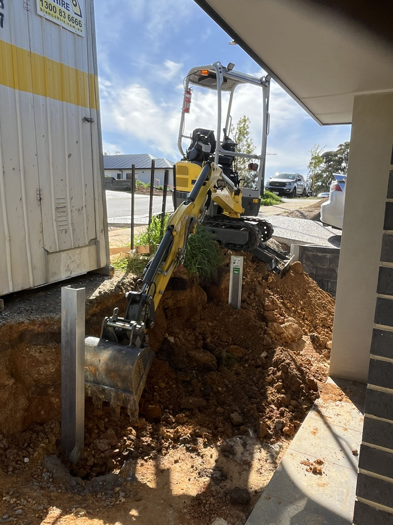 Mini excavator digging beside a new house, with a steel post standing in the hole