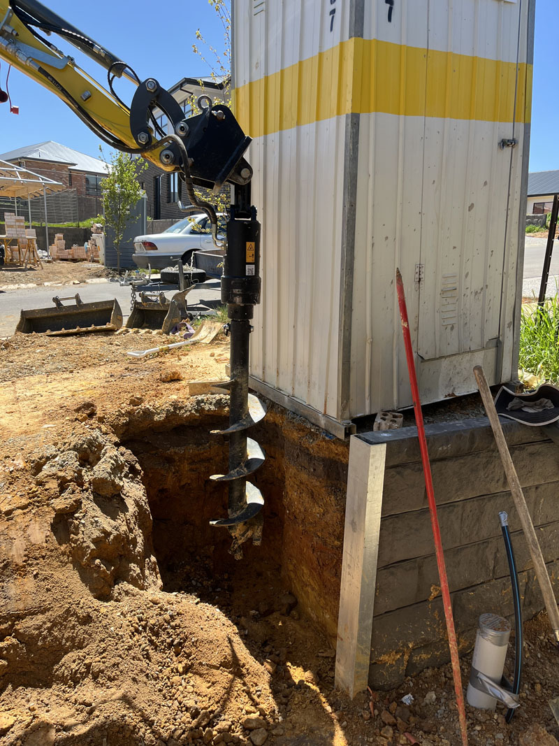 Auger attachment drilling into an excavation beside a portable site cabin