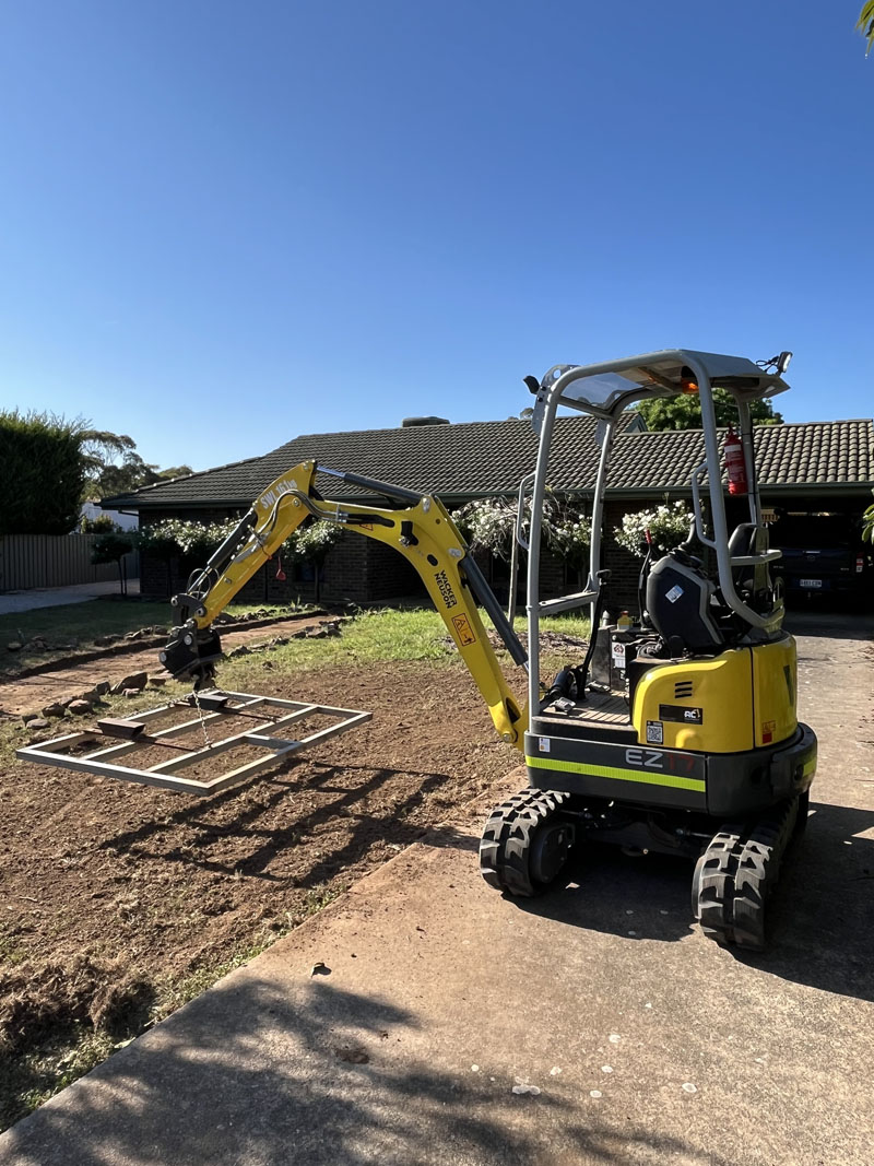 Mini excavator with a levelling frame grading a front yard beside a driveway