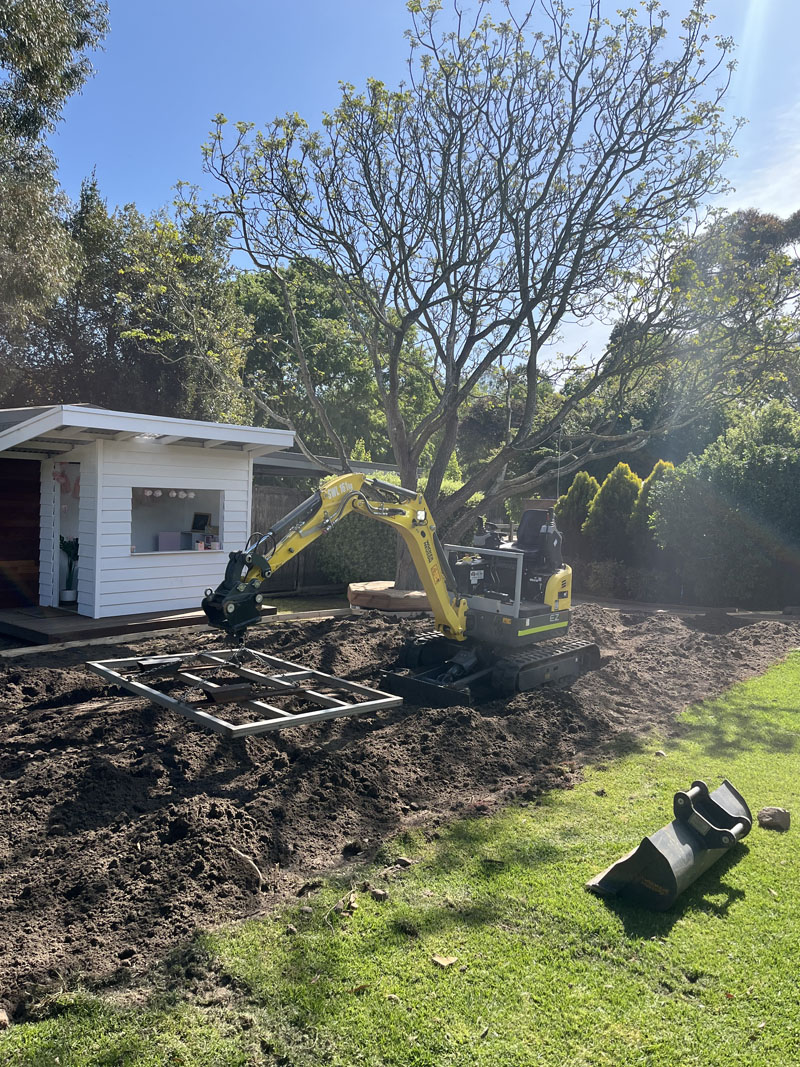 Mini excavator levelling soil beside a white garden shed, with a spare bucket on the lawn