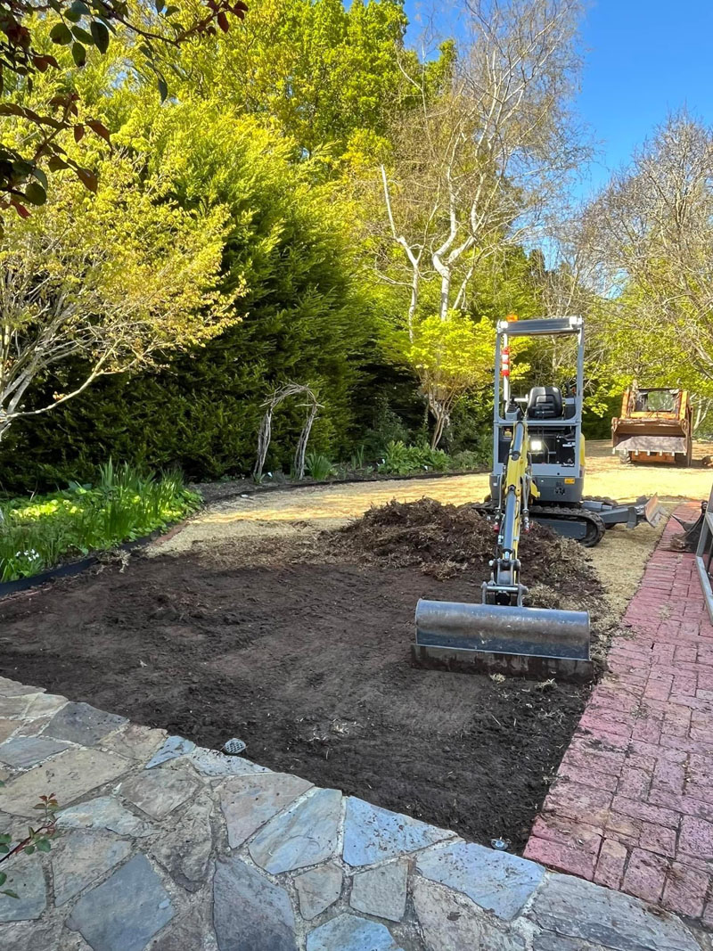 Mini excavator levelling a garden bed beside a brick path and stone paving
