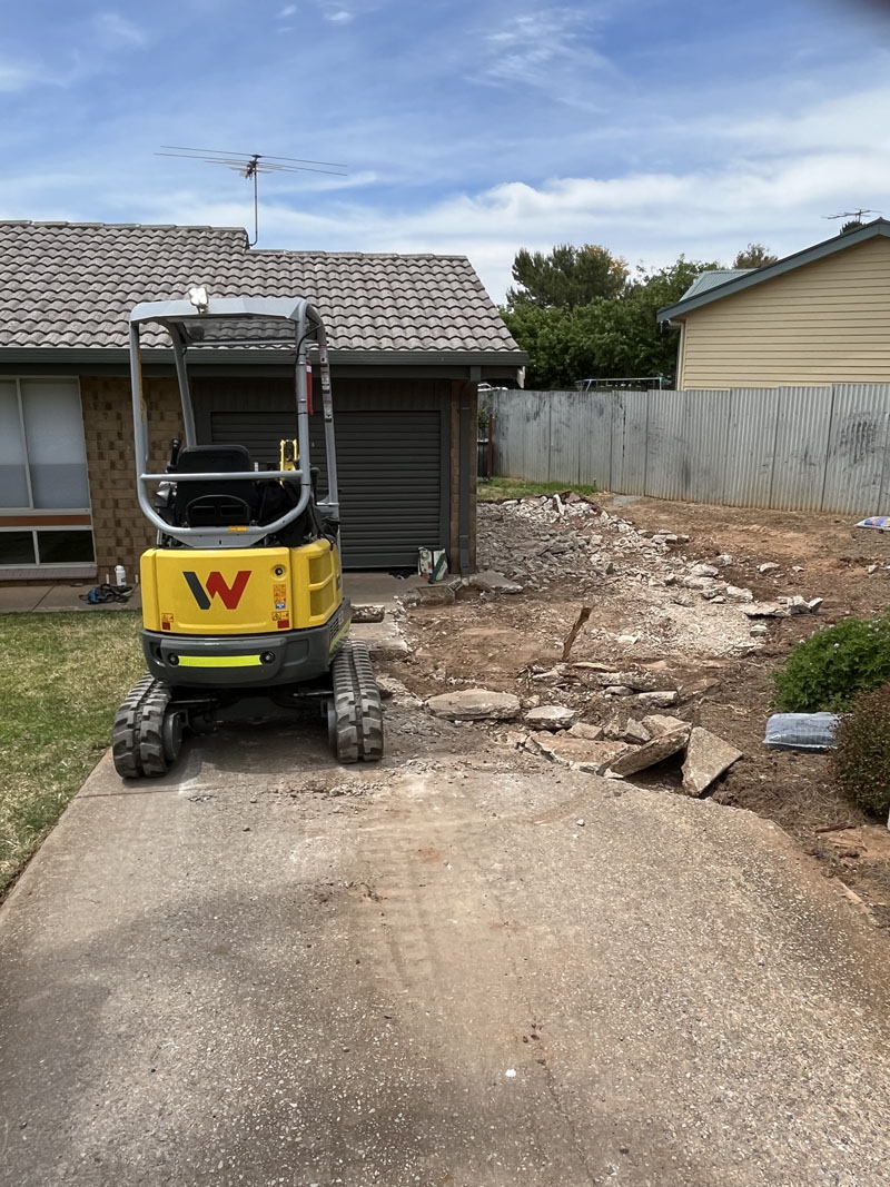 Mini excavator on a driveway beside a front yard strewn with broken concrete