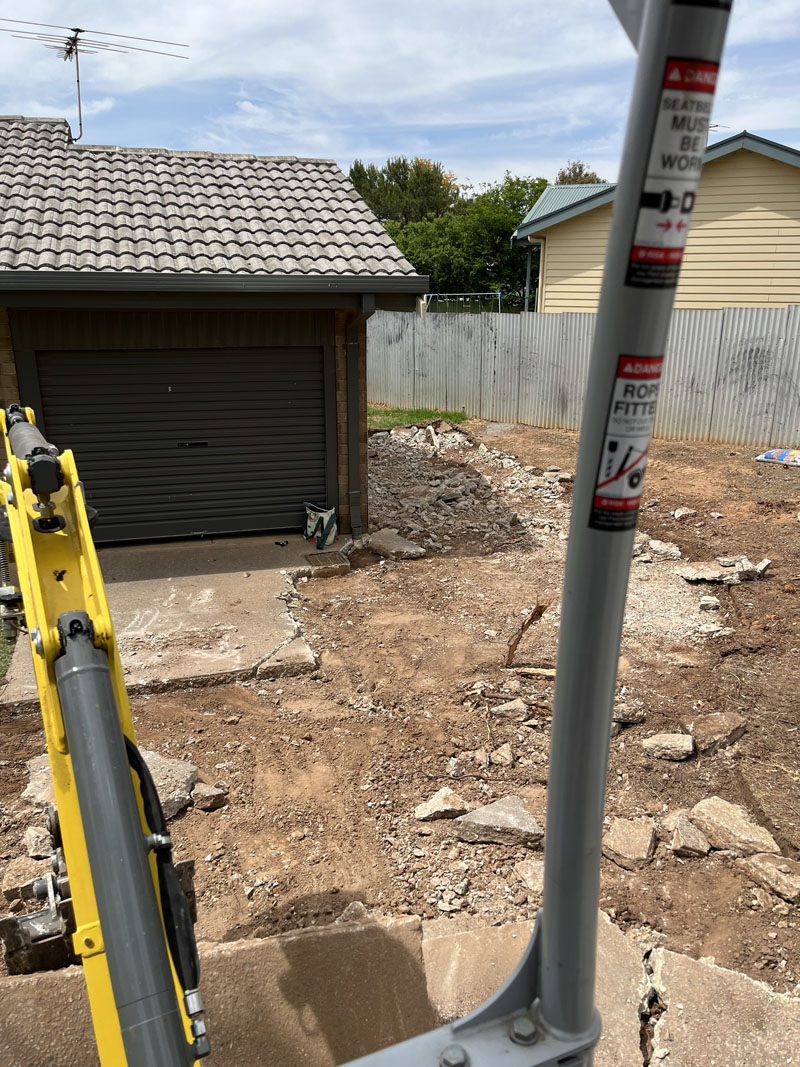 Broken concrete and rubble across a front yard, seen past the excavator's arm