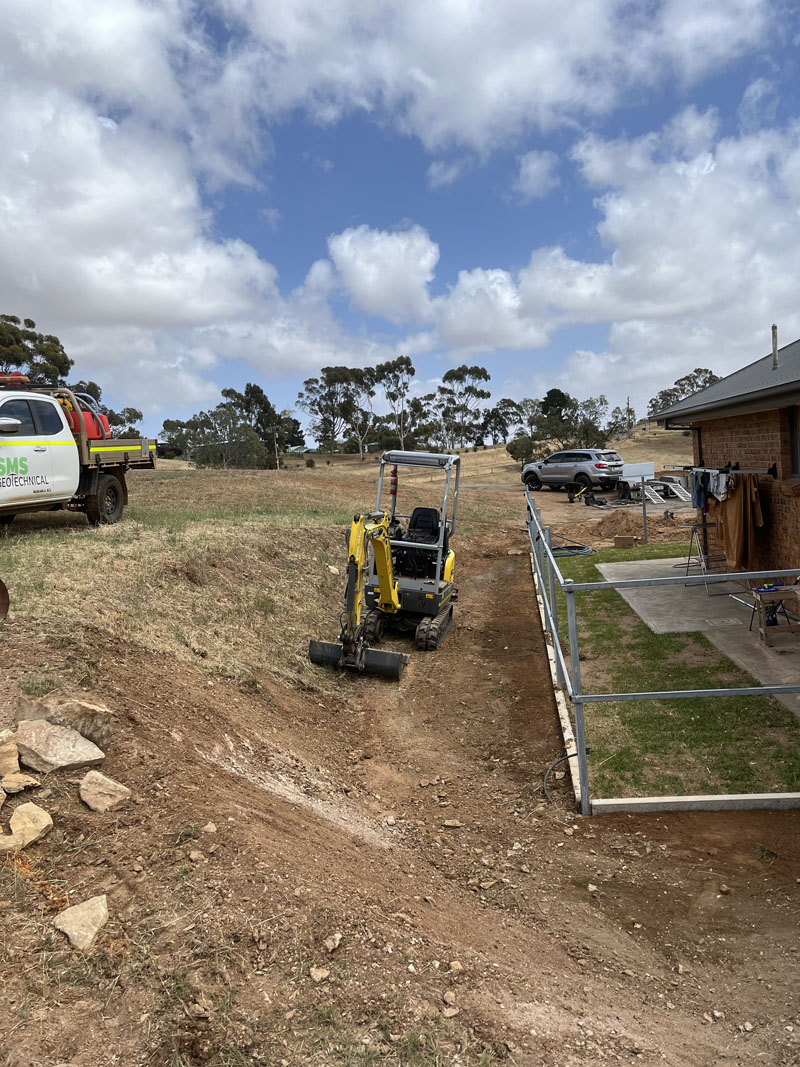 Mini excavator digging a trench down a sloping block beside a fence