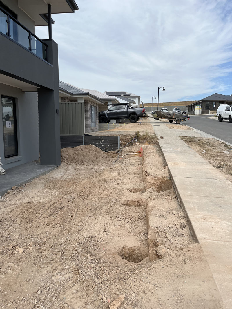 Line of holes dug along the edge of a footpath beside a new two-storey house