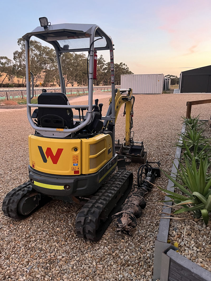 Yellow mini excavator with an auger attachment parked on gravel at sunset