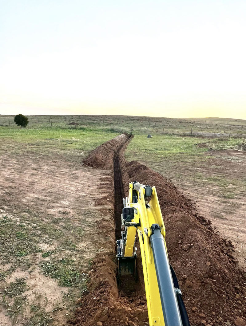 Excavator digging a long, narrow trench across an open paddock