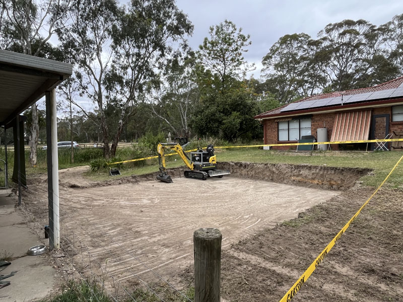 Large levelled site cut taped off beside a brick house, mini excavator behind
