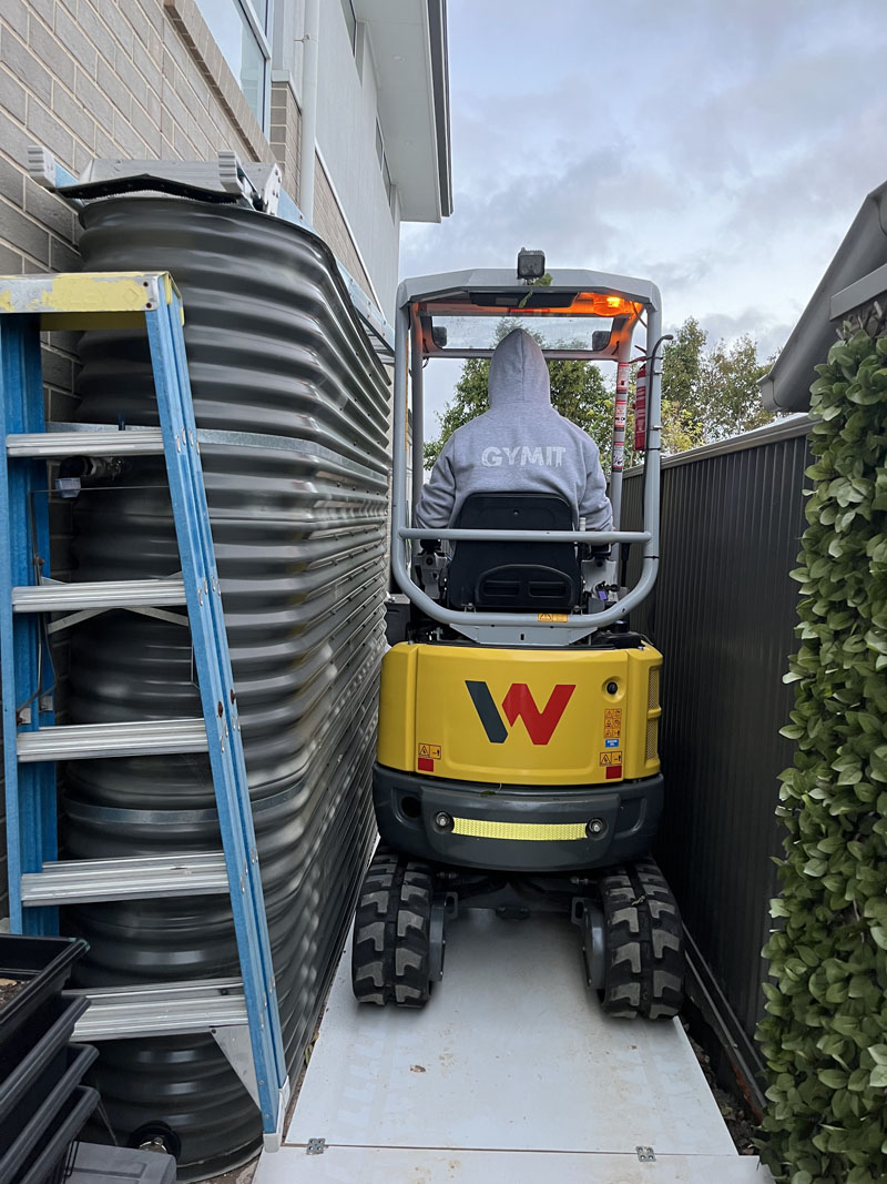 Mini excavator in a narrow side passage between a water tank and a fence