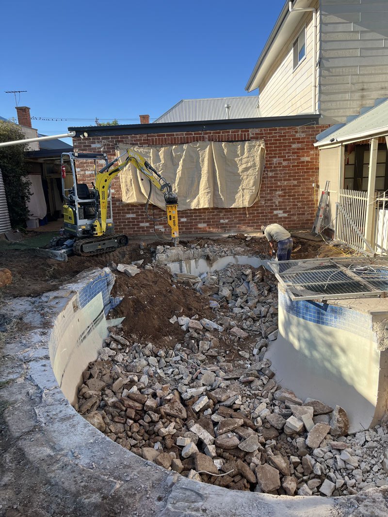 Old in-ground pool shell being filled with broken concrete, mini excavator behind