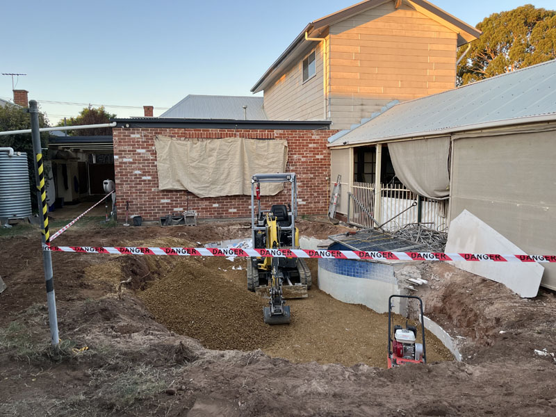 Old pool area being filled behind danger tape, with a mini excavator and plate compactor