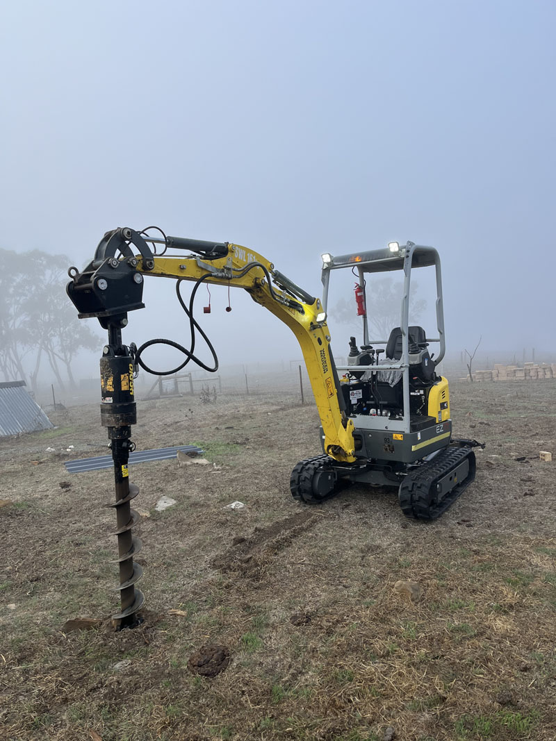 Mini excavator drilling a post hole with an auger in a foggy paddock